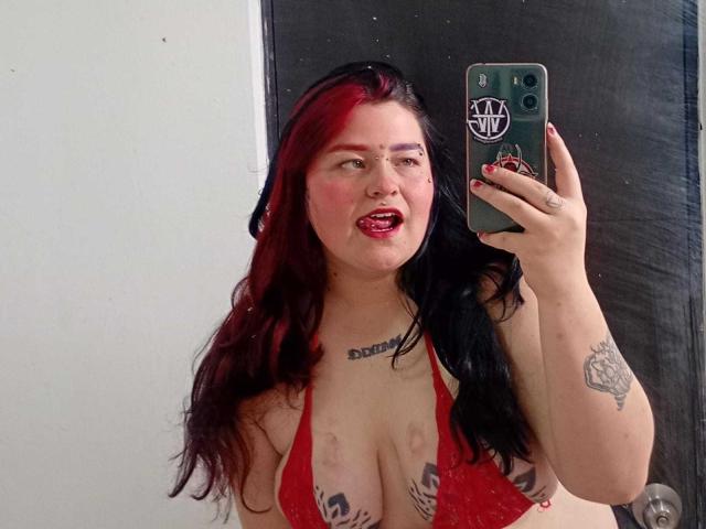 MilaDallila, avec des cheveux roux et noirs, la langue tirée, portant un soutien-gorge en dentelle rouge, montrant ses seins avec des tatouages, tenant un téléphone, selfie devant un miroir, tatouages sur la poitrine et le bras..