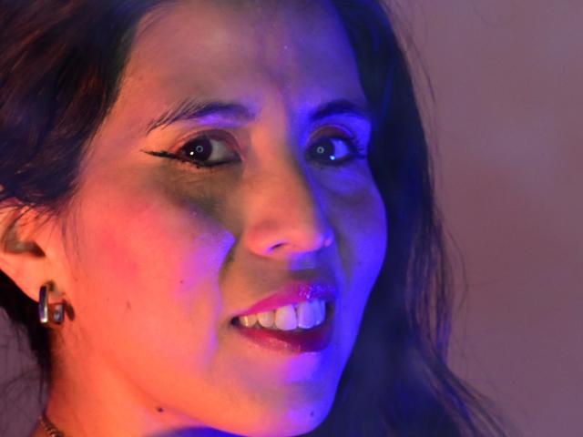 El rostro de LizzHosk con luz azul y rosa sobre su piel, cabello oscuro, delineador de ojos negro, lápiz labial rojo y pequeños aretes, sonriendo mostrando los dientes..