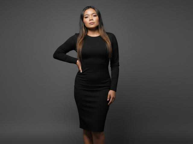 XinemaBonet has long brown hair, wearing a tight black long-sleeve dress, standing with one hand on her hip, showing her ass and small tits. Dark gray background.