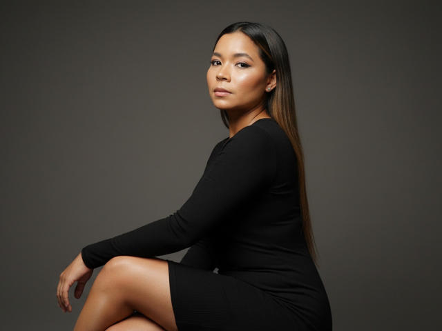XinemaBonet with long brown hair, wearing a tight black dress, sitting with legs crossed, looking at camera, soft brown skin, subtle makeup, confident expression, dark gray background.