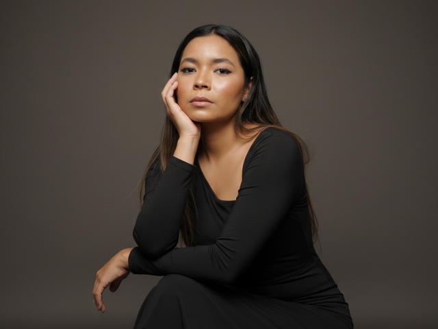 XinemaBonet with long black hair, wearing a black long-sleeve dress, sitting with one hand on her cheek, looking slightly bored, soft brown skin, small nose, subtle makeup.
