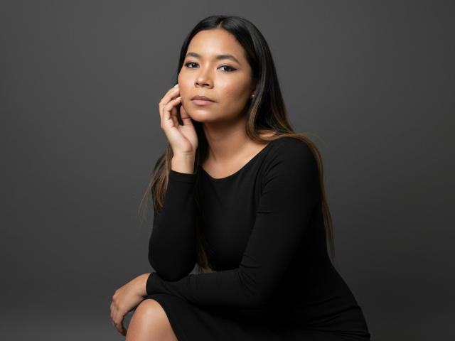 XinemaBonet, Asian woman with long black hair, wearing a black long-sleeve dress, sits with hand on cheek, looking serious, soft brown eyes, slight smile, grey background.