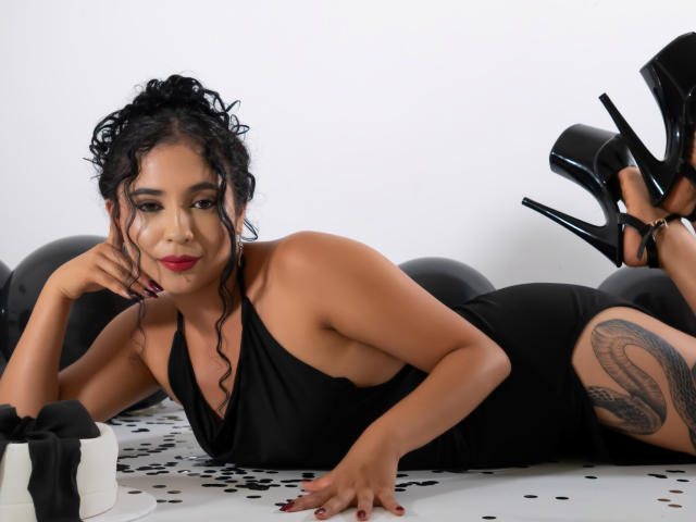 LilithStygian laying on her stomach, wearing a black halter dress, black high heels, and showing a tattoo on her right thigh. Boobs partially visible, black curly hair.