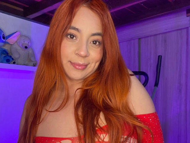 CereScarlett with red hair, pink lips, and a smile, wearing a red off-shoulder top, showing her tits, in a purple-lit room with a teddy bear.