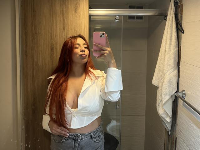 CereScarlett with long red hair, pouty lips, wearing a white shirt exposing her tits, and gray jeans, taking a mirror selfie in a bathroom, holding a pink phone.