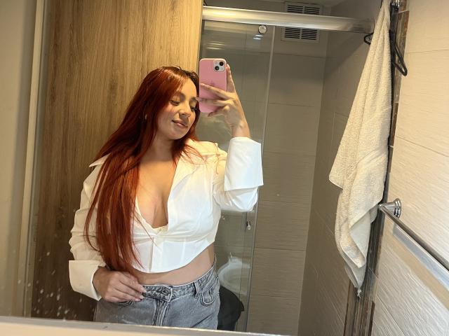 CereScarlett with long red hair, white open shirt showing her big boobs, and gray jeans, taking a mirror selfie with a pink phone in a bathroom.