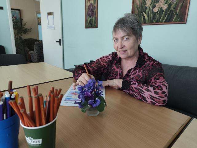 OlgaLuck, middle-aged woman with short gray hair, wearing a pink leopard print shirt, sitting at a wooden table, holding a purple flower vase, writing with an orange pencil.