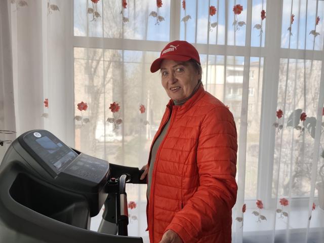 Olga, com boné vermelho e jaqueta vermelha volumosa, em pé em uma esteira, sorrindo, vestindo uma camisa cinza, olhando por cima do ombro, porte médio, pele clara, cabelo castanho curto, ambiente interno..