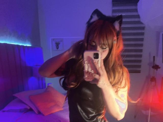 AmaraKazumi with cat ears, long red-orange hair, and black leather top, taking a selfie in a dimly lit room, showing her tits and holding a phone with a pink case.