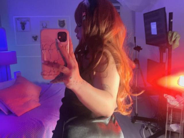AmaraKazumi with long red hair taking a selfie, showing her side profile with a tattoo on her arm, holding a pink phone, in a dimly lit room.