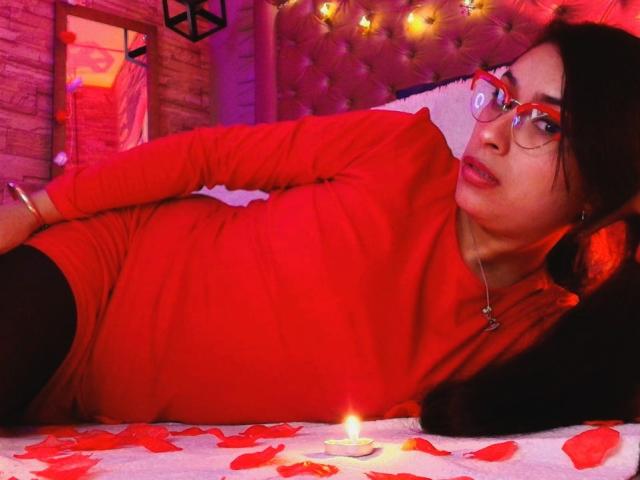 RoxanneHan lying on a bed in a red long-sleeve top, black pants, red glasses, and a necklace. Red rose petals and a lit candle are on the bed.