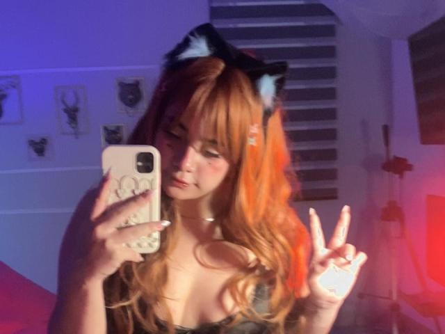AmaraKazumi with orange hair and black cat ears, taking a selfie in a mirror, making a peace sign, showing her tits, wearing a choker, in a dimly lit room.