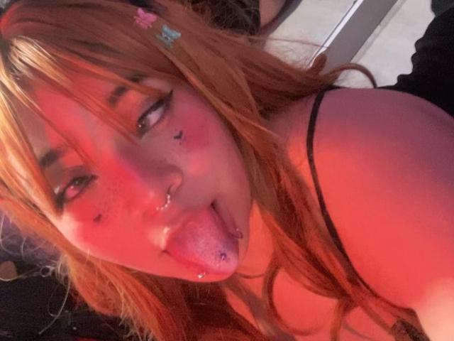 AmaraKazumi has long orange hair, a nose ring, and a tongue piercing. She's sticking out her tongue, looking at the camera with red lighting on her face.