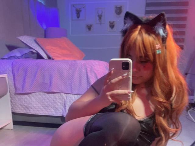 AmaraKazumi with orange hair, cat ears, black top, and thigh-highs, taking a mirror selfie, showing her tits, in a bedroom with a bed and blue lighting.
