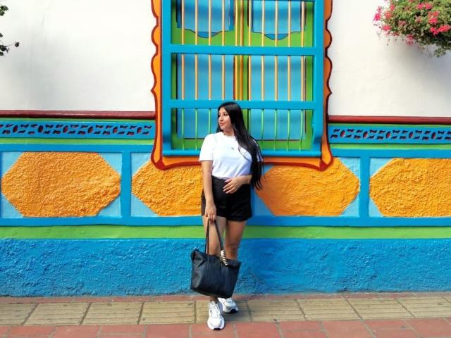 Catalina Rivero, con cabello negro largo, camiseta blanca, pantalones cortos negros, sosteniendo un bolso negro, de pie frente a una pared de colores, con patrones de corazones naranja y azul, usando zapatillas blancas..