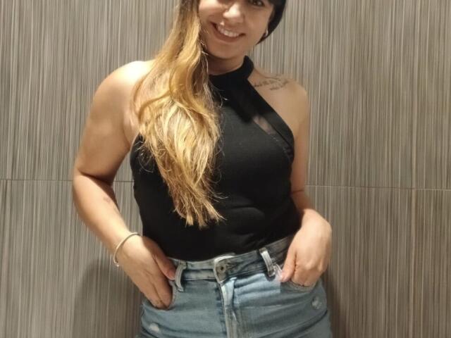 Roxanne Blair, con cabello rubio largo, un top de tirantes negros y jeans azules, sonriendo, con las manos en los bolsillos, mostrando un pequeño tatuaje en su hombro derecho. Lleva puesto un gorro negro..