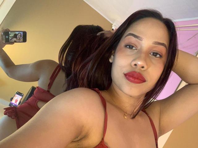 SusanBloom met rode lippenstift, donkerbruin haar en een rode jurk, die een telefoon vasthoudt in een spiegelzelfportret, waarbij haar borsten en oksel in de reflectie te zien zijn. De spiegel laat ook haar rug zien..