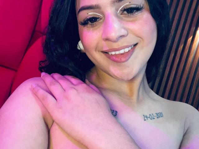 LonxEva with dark hair, smiling, wearing white earbuds, crossed arms, small tattoo on right chest, naked, pink and purple lighting, pink upholstered background.