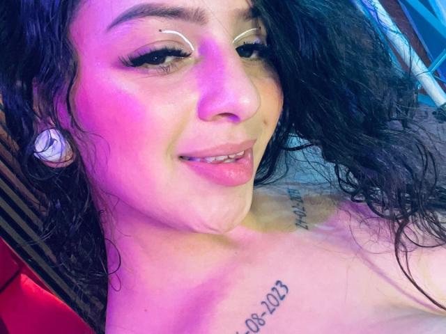 LonxEva with wet black hair, pink and purple lighting, smiling, white eyebrows, nose piercing, tattoos on chest, and visible tongue, wearing large white earbuds, topless.