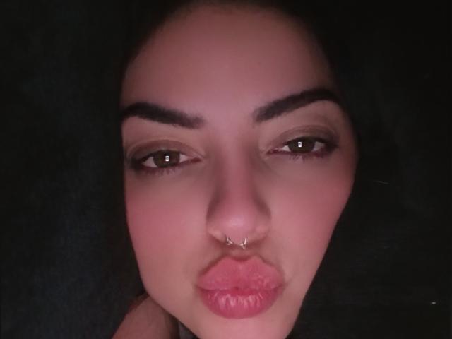 LilithAmoth's face with dark eyebrows, brown eyes, and a small nose piercing. She's pouting with pink, slightly glossy lips. Dark background, slight pinkish-red lighting on her skin.