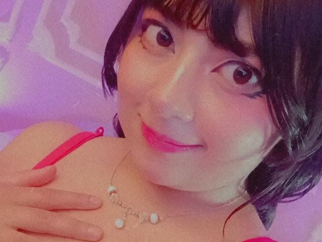 NekoPam has short black hair, pink lipstick, and a cute smile. She's wearing a red top with a necklace and touching her chest with one hand.