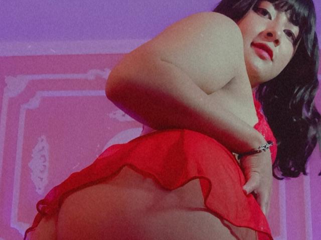 NekoPam with black hair, red lipstick, and red lingerie, showing her ass while looking down at the camera. Her hand is on her butt. Purple light.