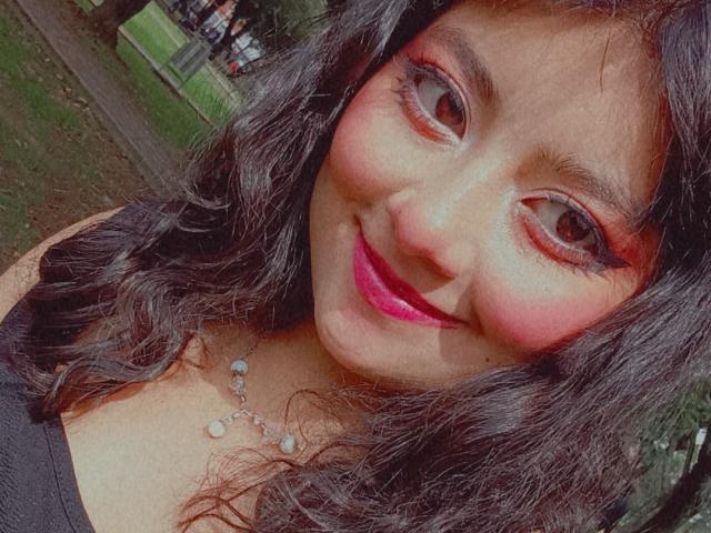 NekoPam has dark wavy hair, pinkish-red eyeshadow, and bright pink lipstick. She's smiling with pink lips, wearing a black top and a necklace with small pearls.