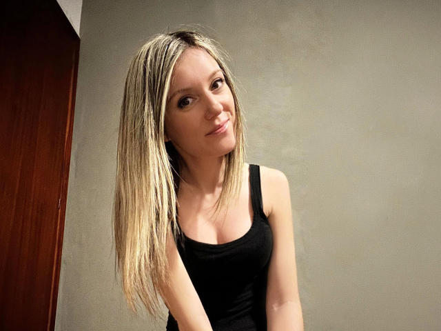 HeisseGina-hot with long blonde hair, wearing a black tank top, slight smile, standing against a plain wall, brown wooden door on the left.