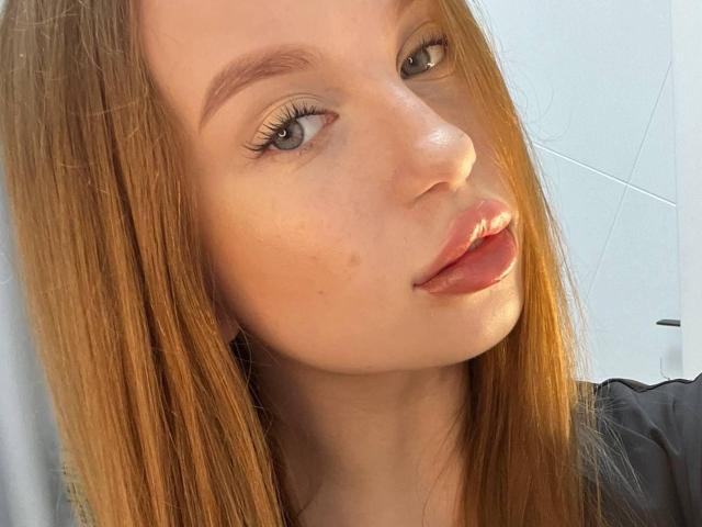 BellaLuvv's face with light brown hair, blue eyes, and glossy pink lips. She's looking slightly to the side with a subtle smile, showing a small zit on her cheek.