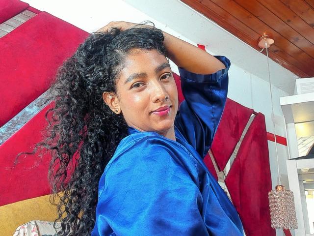 NatalyBlonder with curly black hair, wearing a shiny blue robe, has a cute smile, raised arm, and a small nose ring, in a cozy, warmly lit room.