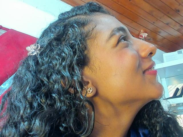 NatalyBlonder's curly black hair, brown skin, and nose ring, looking up with a slight smile, red lipstick, small earrings, wooden ceiling, and a red and white background.