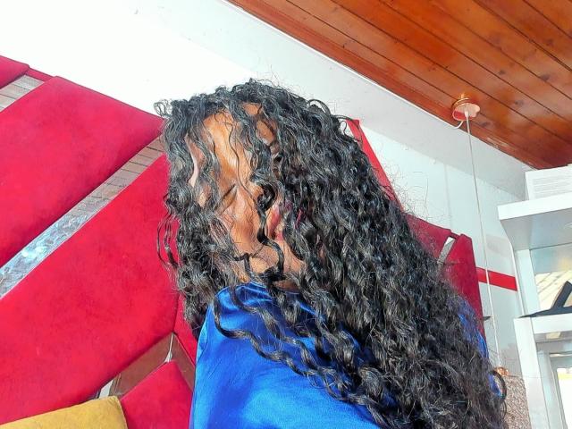 NatalyBlonder with curly black hair, blue shirt, red wall in background, wooden ceiling, and glass window to the right. Her face partially obscured by her long, wavy hair.