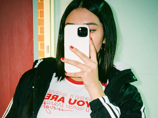 SofiLux with straight black hair, wearing a black and white jacket, holding a white phone covering her face, red nails, white shirt with red text, and a signature.