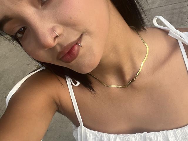 SofiLux has tan skin, a nose ring, and a lip ring. She's wearing a white dress with thin straps and a gold chain necklace. Her boobs are partially visible.