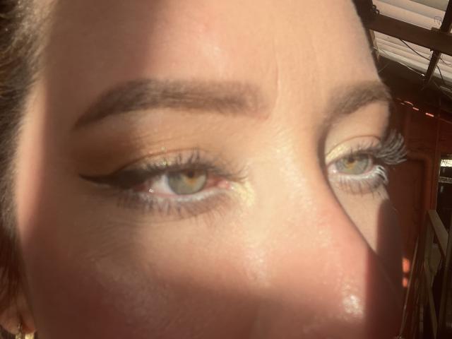 JennyLawry's face with shiny eyes, thick eyebrows, and yellow-green irises, wearing subtle yellow eyeshadow and long eyelashes, in sunlight. Boobs not visible, focus on eyes and brow.