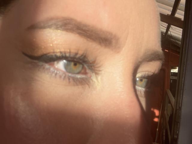 JennyLawry's eye, reflection, and eyebrow, with shimmer eyeshadow, black eyeliner, and long eyelashes, in soft sunlight. Her blue eye and slightly flushed cheek are visible.