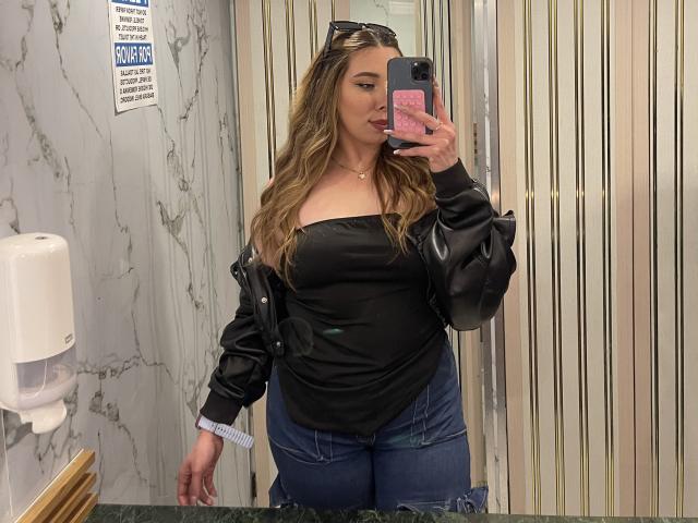 JennyLawry with long blonde hair, black strapless top, black leather jacket, blue jeans, holding phone in mirror, slightly smiling, wearing sunglasses on head.