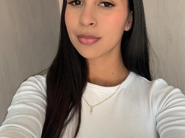 HalleySexy a de longs cheveux noirs, une peau brun clair et des lèvres roses. Elle porte une chemise blanche et un collier avec une croix dorée, avec un sourire discret et de petites boucles d'oreilles..