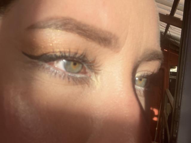 JennyLawry's eye shot, looking through a window, showing her blue eye with thick black eyeliner, glossy eyeshadow, and well-defined eyebrows, with sunlight casting a shadow on her cheek.