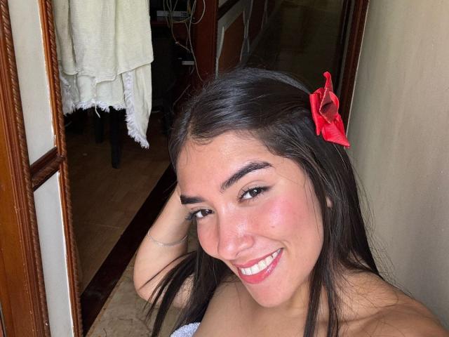 SharonKlart has dark brown hair with a red bow, thick eyebrows, and a big smile showing white teeth. She's topless, with visible pink cheeks, and a cute face.