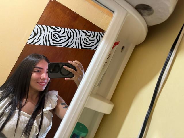 SharonKlart with long black hair, smiling in mirror selfie, holding phone, wearing white ruffled shirt, small tattoo on arm, yellow wall, zebra-patterned wood, toilet paper roll.