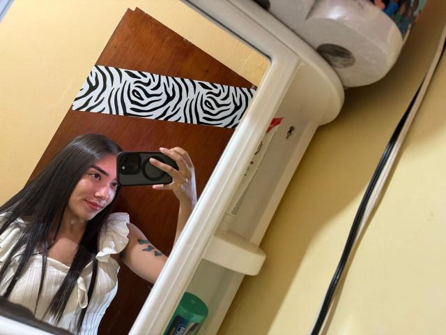 SharonKlart with long black hair, small tits, and tattoos, taking a mirror selfie with a black phone, wearing a white top, in a yellow-walled room.