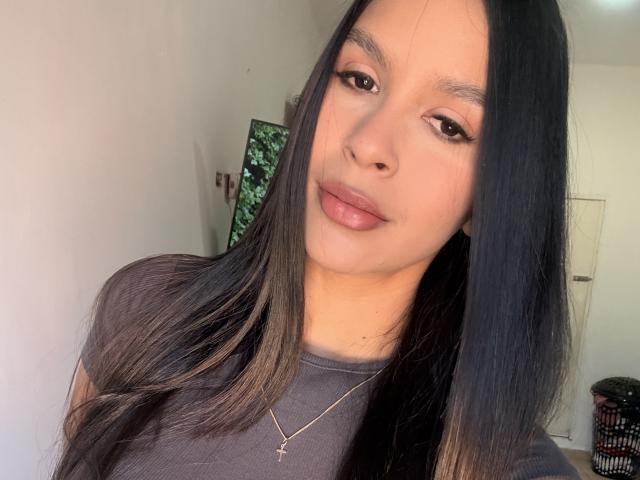 HalleySexy has long black hair, brown eyes, and full pink lips. She's wearing a grey top with a gold cross necklace, and she's looking at the camera with a slight smile.