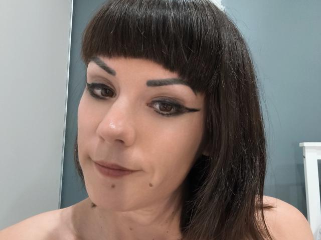 OrianaLaFrancaise has dark brown hair with bangs, black eyeliner wings, and a small beauty mark under her right eye. She's smiling slightly with her bare shoulders visible.