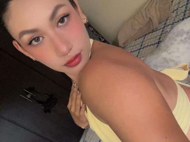 EmilyRobertts has a nose ring, dark eyebrows, red lipstick, and is wearing a yellow top, showing her wet shoulder and back. She's looking at the camera with a slight smile.