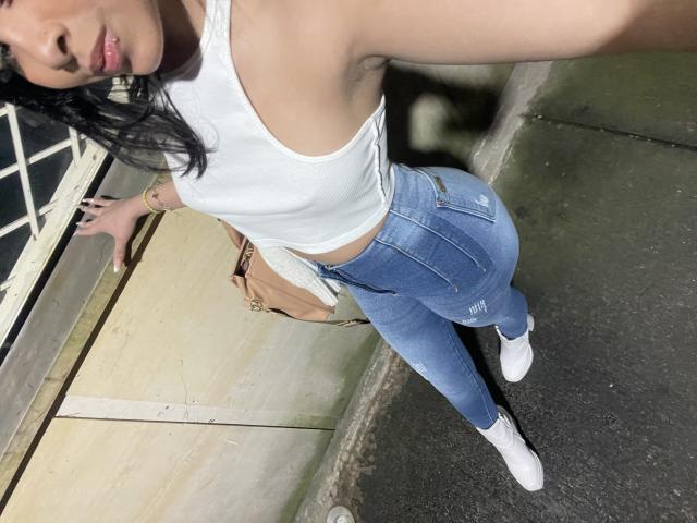 ChloeVixen with medium tits in a white tank top, blue high-waisted jeans, and white socks, standing with one hand on a railing, black hair, and a tan bag.