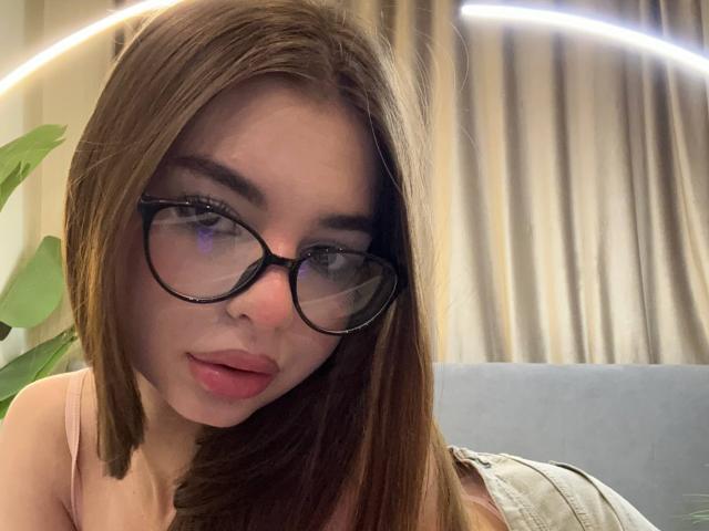 RoseleeVandiest with brown hair, black glasses, pink lipstick, and a cute face, wearing a pink top, leaning forward, showing a subtle pout. She has small, perky tits.
