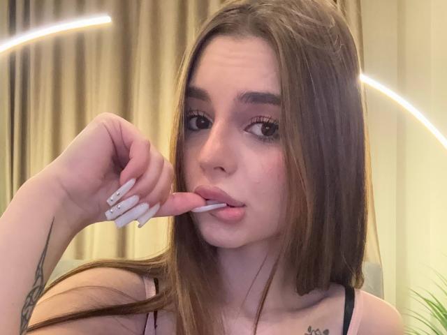 RoseleeVandiest with long brown hair, biting pink lipstick off white nail with long white nails, looking at camera, wearing black strap, tattoo on left arm.
