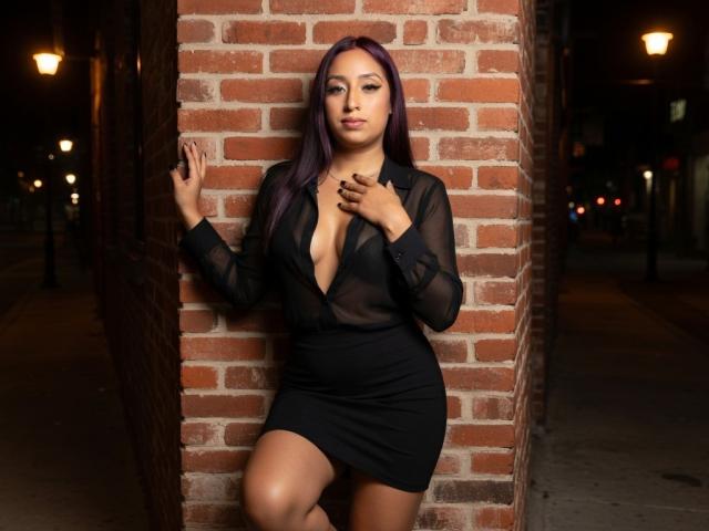 Roxanne Marquez, con i capelli viola, indossa una maglia nera trasparente a maniche lunghe, con un profondo scollo, una gonna nera e si appoggia a un muro di mattoni. È notte, le mani sono appoggiate al muro e sullo sfondo si vedono i lampioni..