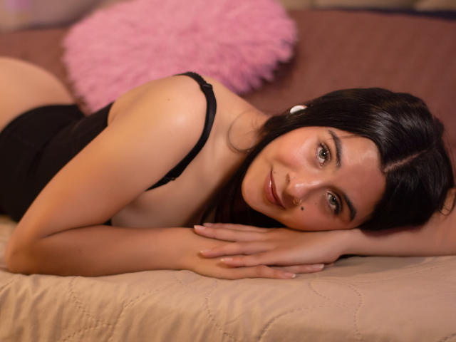 ChloeXk, with dark hair and a cute nose piercing, lies on a bed in black lingerie, smiling at the camera, her hands under her chin, showcasing her small ass.
